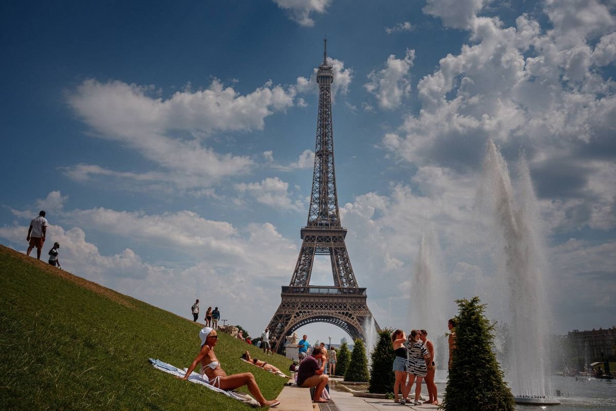 រូបភាពបង្ហាញពីប៉ម Eiffel ដែលជាកន្លែងសម្គាល់មួយ មានទីតាំងនៅក្នុងសួនឧទ្យានមួយដែលមានមនុស្សប្រមូលផ្តុំជុំវិញវា។ ប៉មនេះត្រូវបានហ៊ុំព័ទ្ធដោយរុក្ខជាតិបៃតង និងលក្ខណៈទឹក ដោយមានពពកមួយផ្នែកដែលអាចមើលឃើញនៅផ្ទៃខាងក្រោយ។
