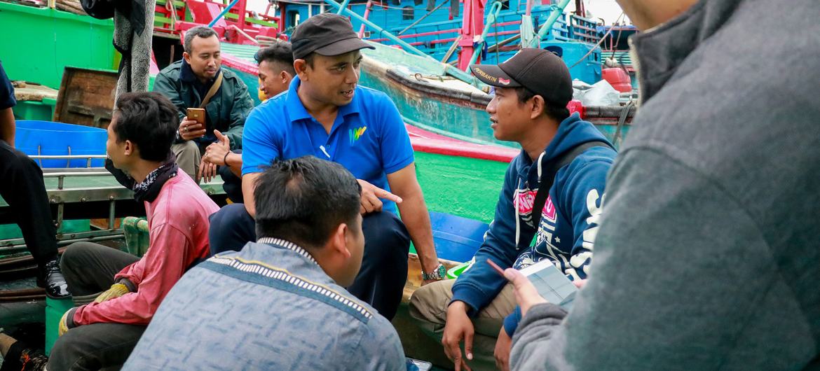 មន្ត្រី IOM ក្នុងអាវខៀវនិយាយជាមួយពលករចំណាកស្រុកឥណ្ឌូនេស៊ីនៅលើទូកនេសាទក្នុងប្រទេសឥណ្ឌូនេស៊ី។